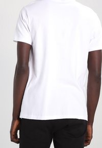 Vit bomulls T-shirt med korta ärmar, rundad hals och rak fåll, sedd bakifrån mot en neutral bakgrund.