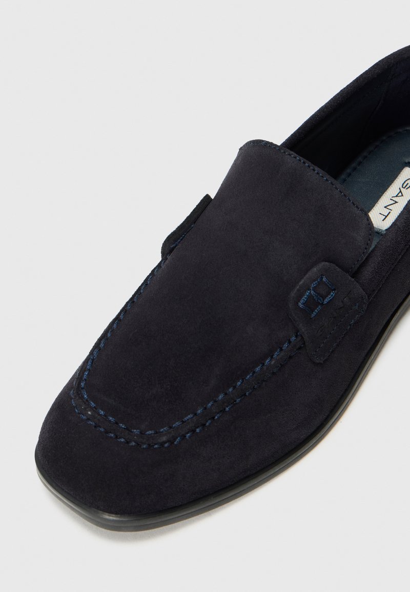 Mocassino in suede blu scuro con punta quadrata, dettagli cuciti e un piccolo emblema cucito sul lato, su sfondo bianco.