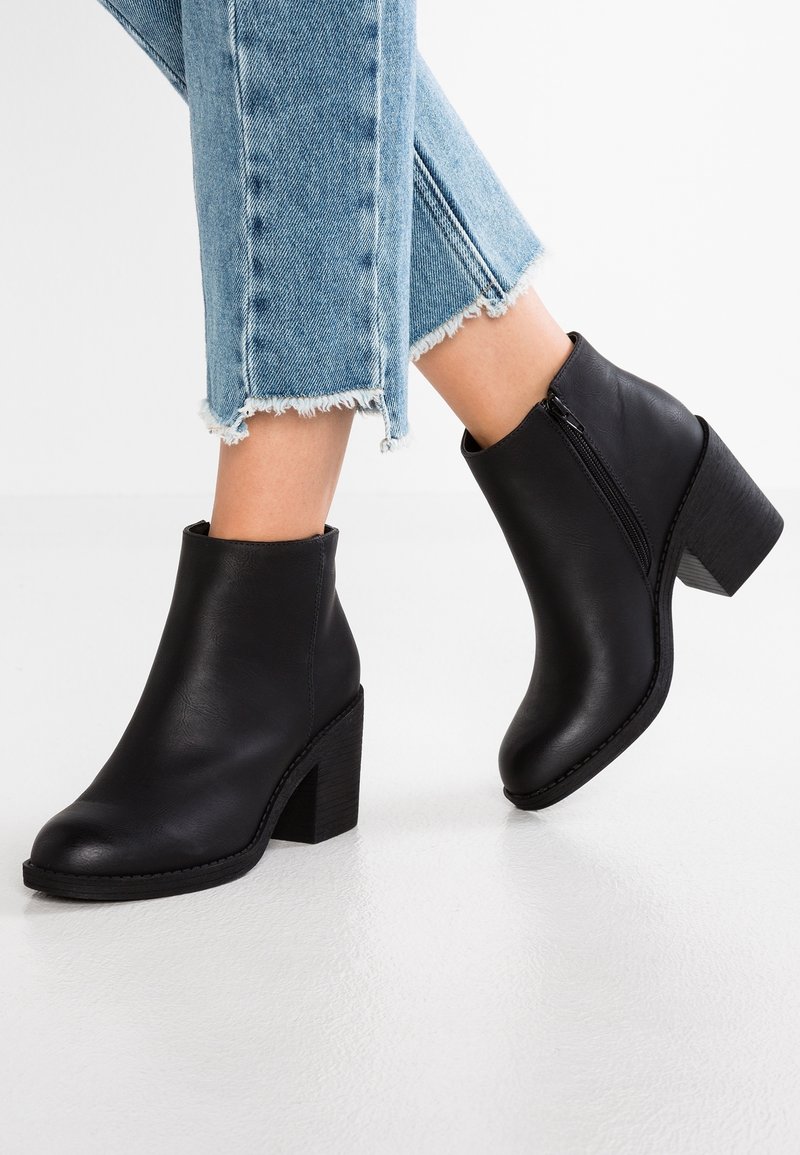 Bottines en cuir noir à talons carrés portées avec un jean court bleu clair à ourlet effiloché sur fond blanc uni.