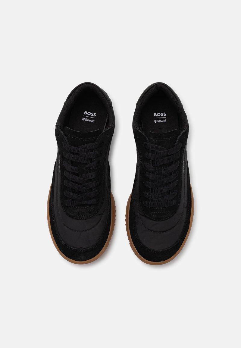 Baskets de sport noires avec une tige en tissu texturé, semelles en caoutchouc et lacets plats noirs. Présente un logo sur la languette et une semelle extérieure marron contrastante.