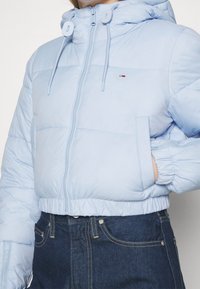 Žena v světle modré zkrácené péřové bundě s kapucí a tmavě modrých kalhotách s vysokým pasem, ruka zasunutá v kapse bundy.