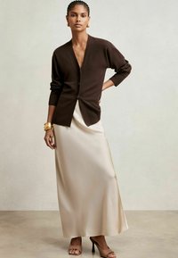 Cárdigan marrón, con cuello en V profundo, puños acanalados, llevado sobre una falda maxi de satén crema. Tacones abiertos en tono beige y una pulsera dorada acentúan el look.