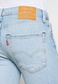 Pantalones vaqueros de mezclilla azul claro con un ajuste skinny y cónico; presentan un parche de cuero con la etiqueta "Levi Strauss & Co." y una pestaña roja en el bolsillo trasero.