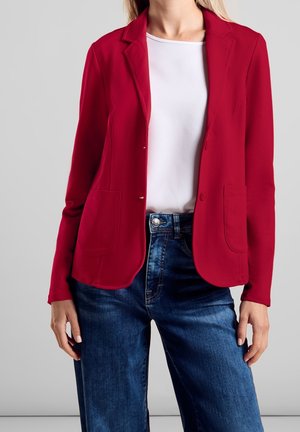 Vrouw met een rood colbert over een witte top en een blauwe spijkerbroek, staand tegen een effen grijze achtergrond.