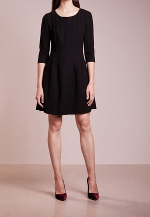 Mujer con un vestido negro hasta la rodilla con mangas tres cuartos y tacones altos burdeos, posando contra un fondo beige liso.