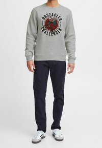 Harmaa collegepaita, jossa on graafinen painatus tekstillä, resorit hihansuissa ja helmassa, yhdistetty tummansinisiin housuihin ja monivärisiin lenkkareihin.