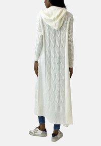 Langer weißer Strickcardigan mit Kapuze, ausgestattet mit einem Zopfmuster und einem gerippten Saum, kombiniert mit blauen Jeans und grauen Sneakers.