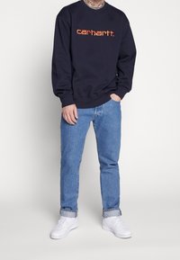 Sudadera navy con logo "carhartt." en naranja, cuello redondo, puños de canalé, combinada con pantalones de mezclilla azul claro y zapatillas blancas.