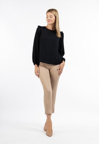 Blusa nera strutturata con dettagli a volant sulle maniche, abbinata a pantaloni beige aderenti e tacchi nudi. Sfondo bianco semplice.