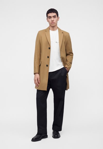 Jeune homme portant un manteau beige sur une chemise blanche et un pantalon noir avec des chaussures noires, debout devant un fond clair uni.