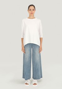 Biała, luźna bluzka z okrągłym dekoltem i rękawami do trzech kvart, zestawiona z szerokimi, jasnoniebieskimi dżinsami i białymi sandałami.