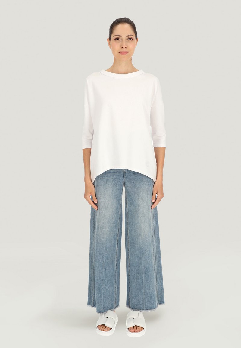 Biała, luźna bluzka z okrągłym dekoltem i rękawami do trzech kvart, zestawiona z szerokimi, jasnoniebieskimi dżinsami i białymi sandałami.