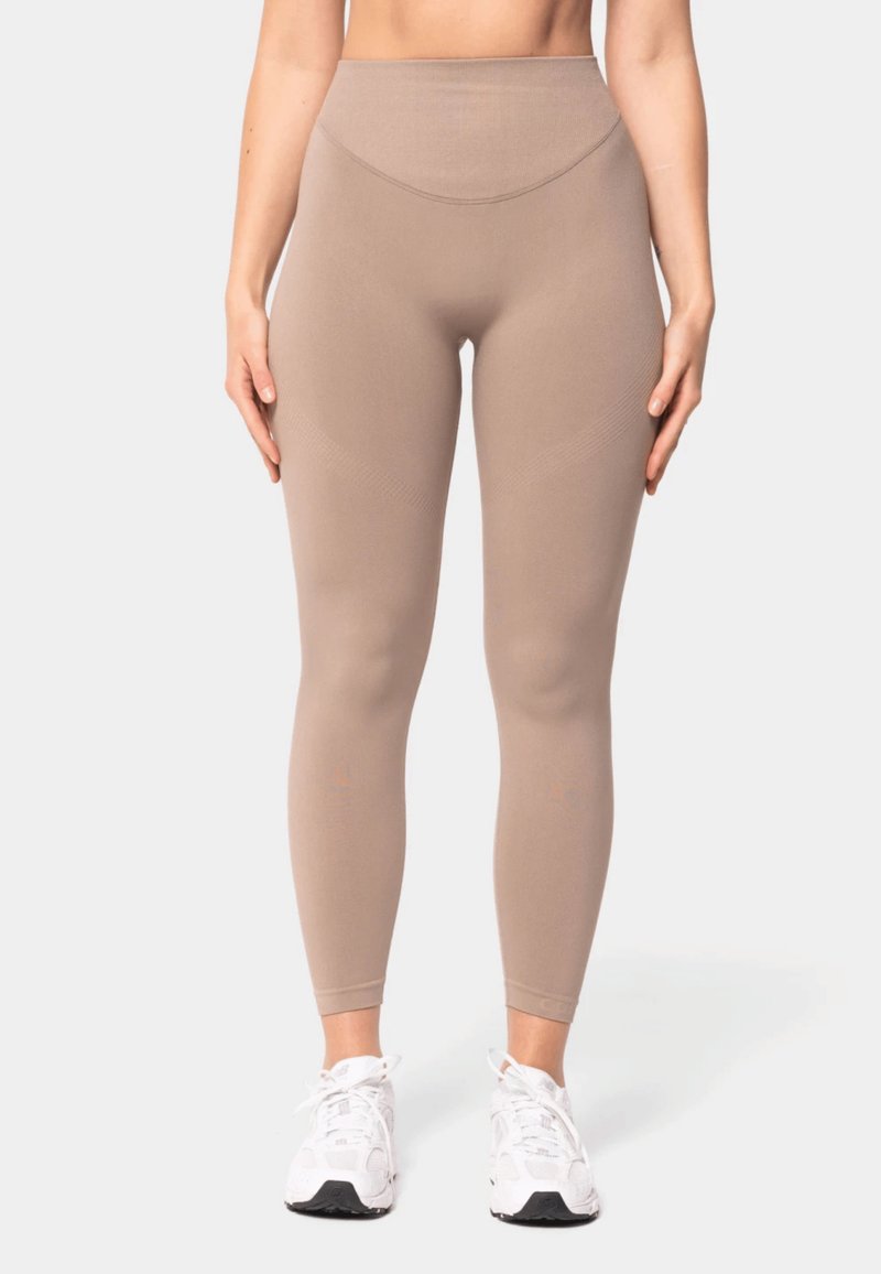 Kvinde iført beige leggings med høj talje og hvide sportssko, der står mod en ensartet lys baggrund.