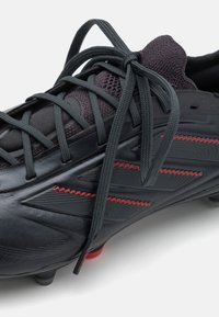 Black football boots with gray laces, featuring directional ridges and red zigzag accents on the upper. Textured synthetic material and molded studs.