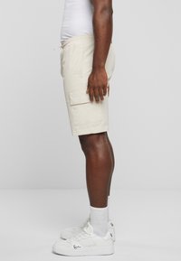 Beige cargo shorts med elastisk midjebånd og sidelommer, med en hvit logoaccent på høyre lomme. Brukt med hvite sneakers.