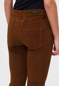 Pantalon en velours côtelé marron avec poches arrière porté par une personne en haut noir, montrant la taille et le haut des jambes de dos.