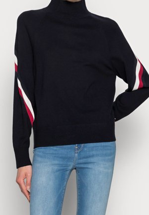 Personne portant un pull à col roulé noir avec des rayures rouges et blanches sur les manches, associé à un jean bleu clair, debout devant un fond uni.