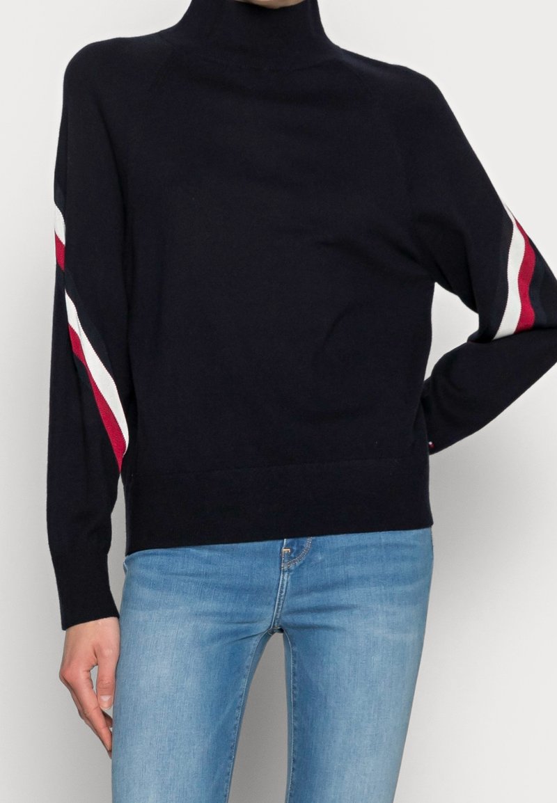 Personne portant un pull à col roulé noir avec des rayures rouges et blanches sur les manches, associé à un jean bleu clair, debout devant un fond uni.