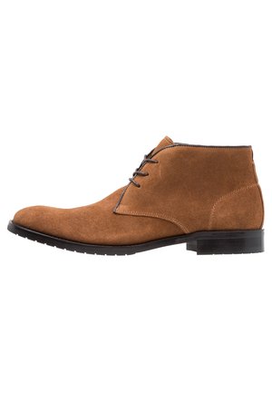 Botines de ante marrón con puntera redondeada, cordones en la parte delantera y una suela de goma texturada baja. Los acentos de costura complementan el diseño.