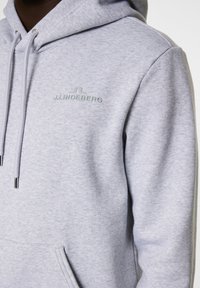 Grå huvtröja med en framficka i kängurustil, dragskohette och präglad "J.LINDEBERG"-logotyp i ljusgrön på bröstet. Mjuk textur.
