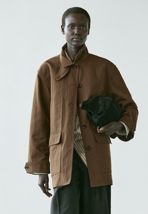 Model draagt een oversized bruine jas met knopen over een gestreept shirt en zwarte broek, houdt een opgevouwen zwart kledingstuk onder één arm.