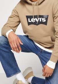 Personne portant un sweat à capuche Levi's beige, un jean bleu, des chaussettes blanches et des baskets beiges, assise avec un genou levé.
