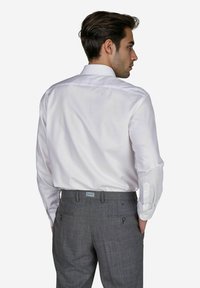Ein Mann steht mit dem Rücken zur Kamera, trägt ein weißes Hemd und graue Hosen und wird vor einem schlichten weißen Hintergrund gesehen.