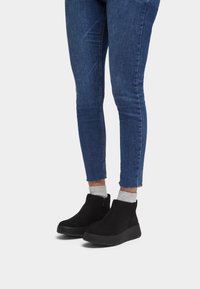 Botas de ante negras hasta el tobillo con un tacón bajo y cremallera lateral, combinadas con unos vaqueros ajustados azules y calcetines grises, mostrando un diseño casual.