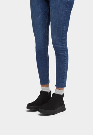 Zwarte suède enkellaarzen met een lage hak en een zijrits, gedragen met blauwe skinny jeans en grijze sokken, wat een casual ontwerp laat zien.