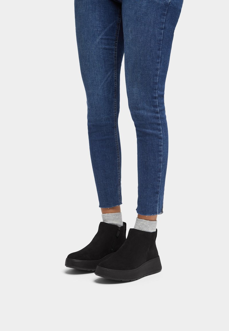 Botas de ante negras hasta el tobillo con un tacón bajo y cremallera lateral, combinadas con unos vaqueros ajustados azules y calcetines grises, mostrando un diseño casual.