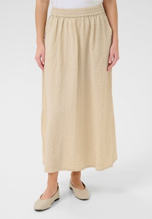 Gonna maxi beige a pieghe con vita elastica, realizzata in tessuto leggero, abbinata a scarpe slip-on beige e una maglietta bianca.