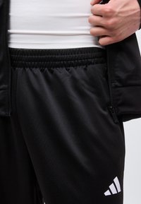 Άτομο που φοράει μαύρο παντελόνι Adidas με ελαστική μέση, λευκό λογότυπο κοντά στο τελείωμα και μαύρο μπουφάν με φερμουάρ πάνω από λευκό πουκάμισο.