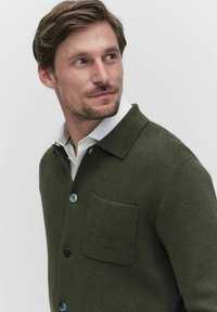 Mörkgrön stickad cardigan med krage, en bröstficka och blå knappar. Tyget har en texturerad yta. Vithalskrage synlig.