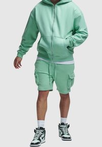 Homme portant un sweat à capuche zippé vert menthe et un short cargo assorti, avec des chaussettes blanches et des baskets montantes noires et blanches.