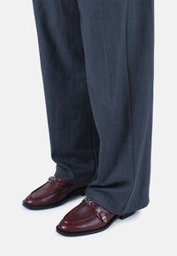Mocasines de cuero marrón con acentos de cadena plateada, combinados con pantalones grises con pliegues. Los zapatos presentan un acabado suave y un diseño de punta cuadrada.