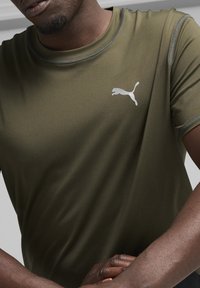 Tamsiai žalia sportinė marškinėlių, pagaminti iš drėgmę sugerdančios medžiagos, turi apvalų iškirpimą ir atspindinčią Puma logotipą kairėje krūtinės pusėje.