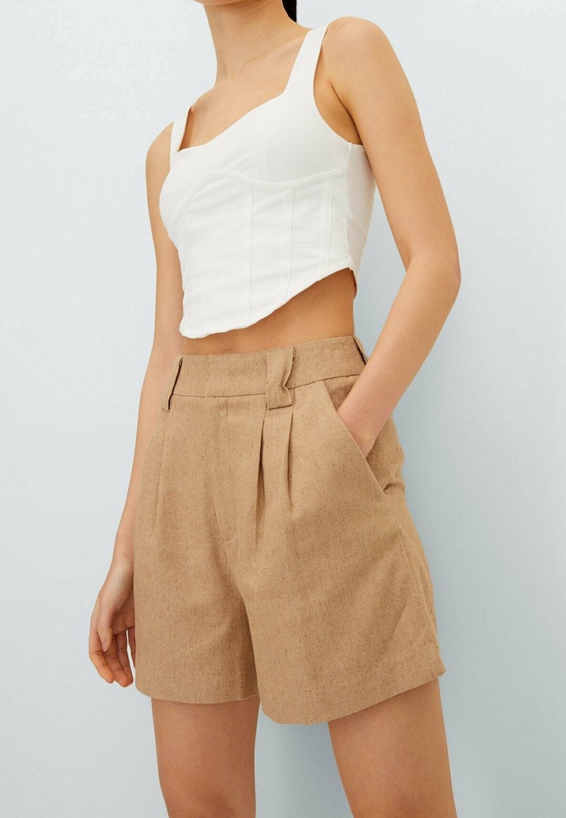 Femme portant un crop top blanc ajusté et un short plissé beige taille haute avec poches, debout devant un fond clair uni.