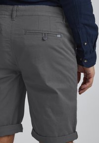 Gray rolled shorts with a smooth texture, featuring back pockets and a button closure. Seen from the side against a plain background.