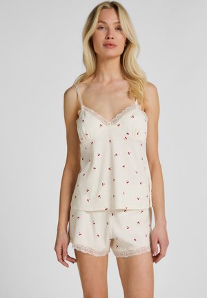 Blondine draag een crèmekleurig topje en korte broekje met rode kersenprint en kanten rand, staand tegen een effen achtergrond.