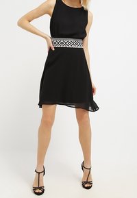Mujer que lleva un vestido negro sin mangas con una cinturilla geométrica en blanco y negro, y sandalias negras de tacón alto con correas en el tobillo.