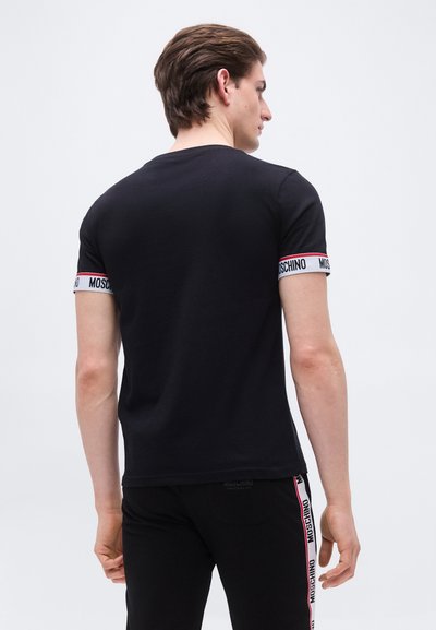 Homme portant un t-shirt Moschino noir et un pantalon avec un bord blanc et rouge de marque, debout de dos sur un fond blanc uni.