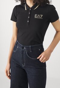 Svart polotröja med gulddetaljer, tre knappar och ribbad krage. Bärs med mörka denimjeans med vit sömnad.