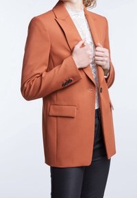 Blazer arancio bruciato con una silhouette leggermente strutturata, chiusura a un bottone, due tasche anteriori e accenti in bottone neri. Texture opaca.