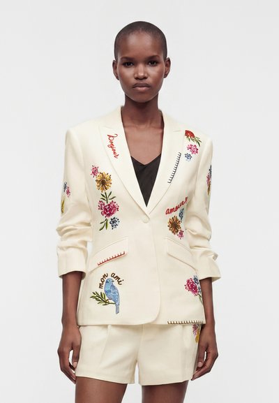 Femme debout, portant un blazer crème et un short avec une broderie florale colorée et des mots en français, sur fond blanc uni.