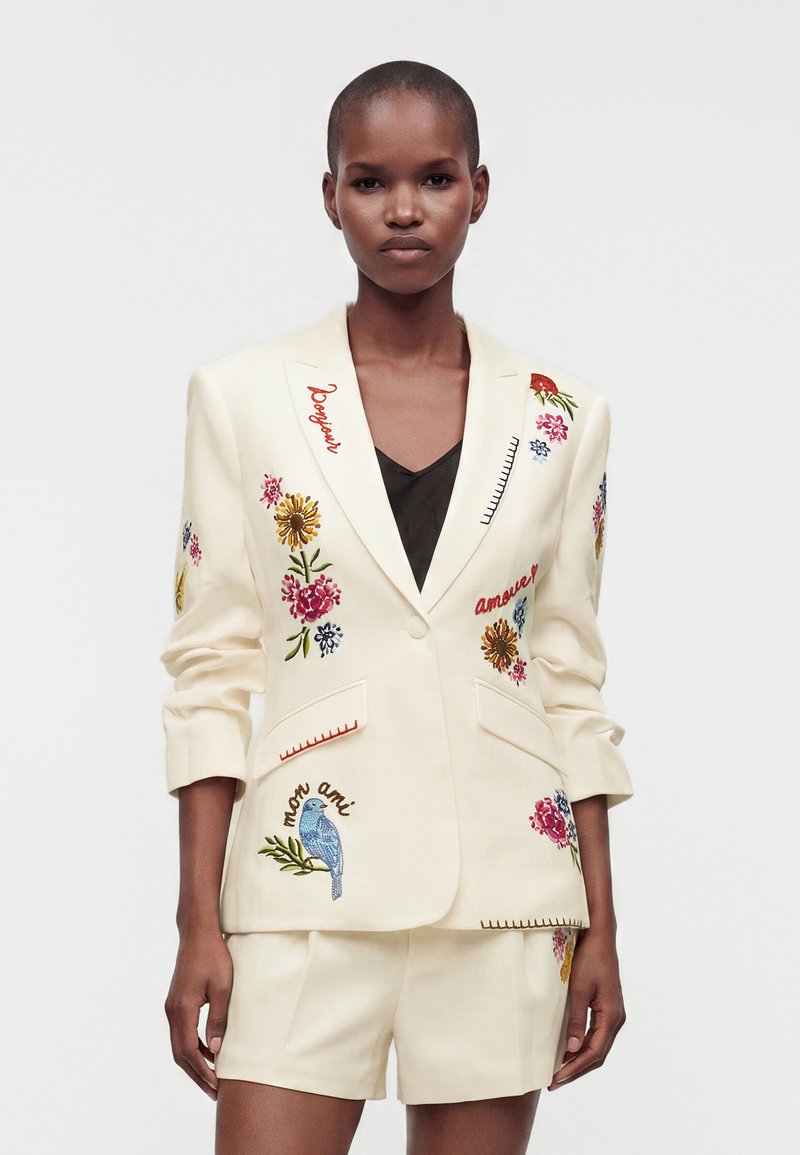Femme debout, portant un blazer crème et un short avec une broderie florale colorée et des mots en français, sur fond blanc uni.
