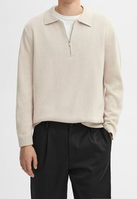 Jasnobeżowy, dziergany sweter z półzamkiem i kołnierzem, noszony na białej koszulce; zestawiony z czarnymi plisowanymi spodniami.
