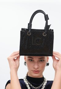 Un mannequin tient un élégant sac à main noir en sequins avec un logo "LC", portant des boucles d'oreilles en argent et un haut en velours avec un collier épais.