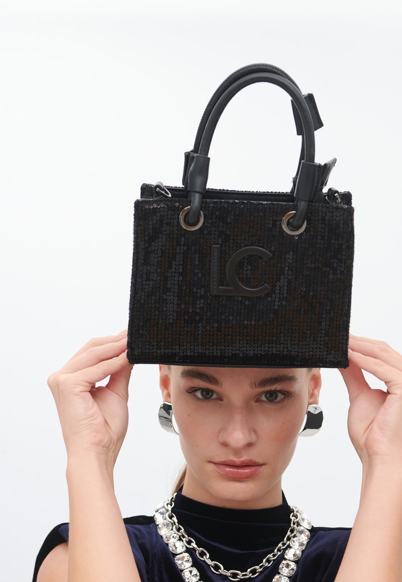 Un mannequin tient un élégant sac à main noir en sequins avec un logo "LC", portant des boucles d'oreilles en argent et un haut en velours avec un collier épais.