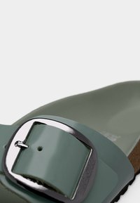 Κοντινό πλάνο ενός πράσινου λουριού σανδαλιού με ένα γυαλιστερό ασημί αγκράφα και μέρος της φελλόσαμπας που φαίνεται κάτω δεξιά.