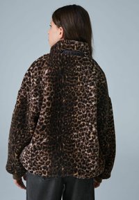 Fleecejacke im Leopardenmotiv mit einem hohen Kragen, weicher Textur, weit geschnittenen Ärmeln und einem Logo auf der Rückseite. Farbpalette umfasst Schwarz- und Brauntöne.